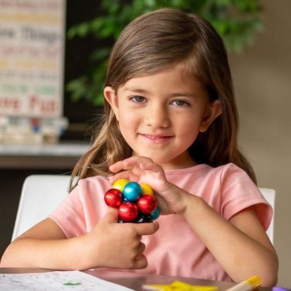 A young girl sits at a table holding a colorful molecular model, with papers and markers in front of her—capturing the spirit of discovery we celebrate at Estrellita En La Casa - About us.